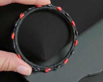 May include: A circular bracelet featuring a black cord base wrapped in a tight, textured pattern. The design is accented with small, evenly spaced bright red loops integrated into the black cord structure, creating a consistent contrast.