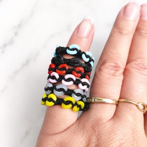May include: A stack of six rings made of black cord woven with small, colorful plastic links in light blue, black, red, pink, translucent white, and yellow. Two additional plain gold-toned metal rings are worn on the adjacent finger.