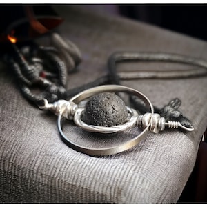 Puede incluir: Un collar de plata con un colgante de piedra de lava negra. El colgante está rodeado por un anillo de plata.
