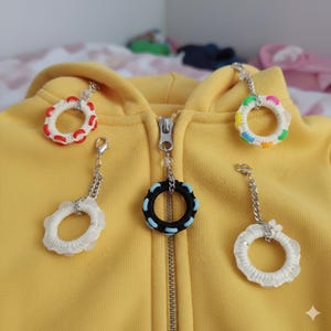 Op de afbeelding: Vijf decoratieve ritstrekkers met cirkelvormige ontwerpen in verschillende kleuren, waaronder rood, wit, zwart en blauw, bevestigd aan een gele hoodie. Elke trekker heeft een zilveren ketting en sluiting.