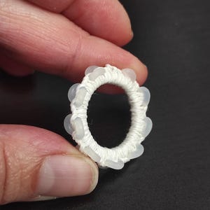 May include: A white, circular handmade ring with small, light-colored beads evenly spaced around the outer edge. The ring is held between fingers against a dark background, showcasing the delicate craftsmanship.