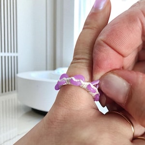 May include: A handmade ring featuring a series of lavender-colored beads woven with white thread. The ring is on a finger, showcasing its design and craftsmanship. The ring's design is simple and elegant.