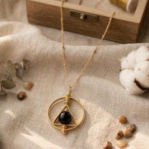 May include: A gold-colored necklace with a circular pendant. Inside the circle is a triangle with a black sphere at its center. The necklace chain is delicate and beaded. The background includes a wooden box, cotton, and small stones.