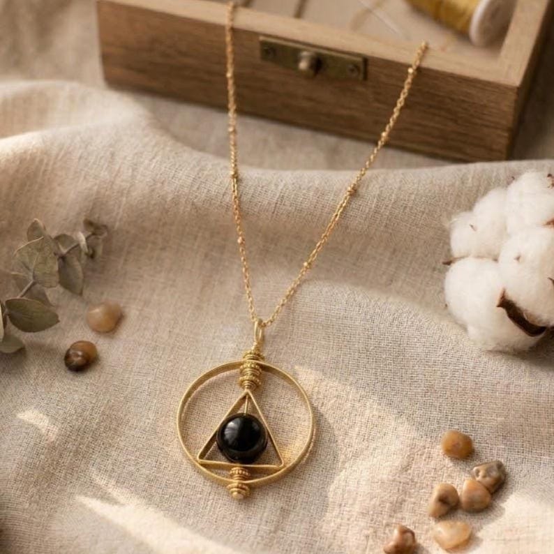 May include: A gold-colored necklace with a circular pendant. Inside the circle is a triangle with a black sphere at its center. The necklace chain is delicate and beaded. The background includes a wooden box, cotton, and small stones.