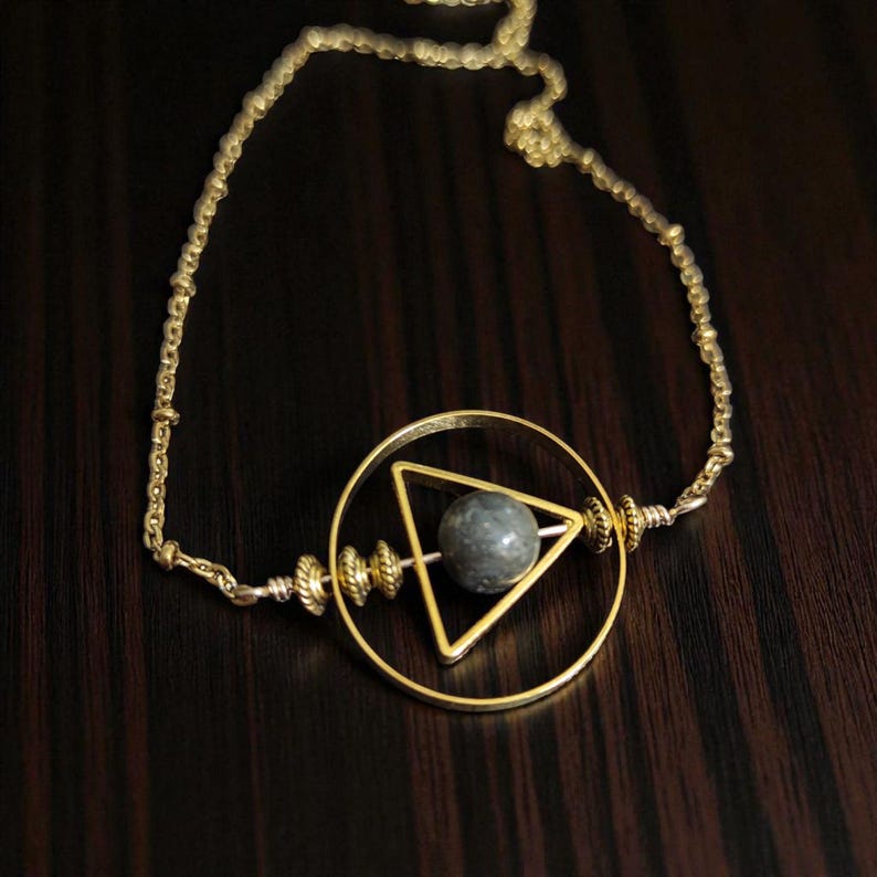 May include: A gold-colored necklace featuring a circular pendant with a triangle and a gray sphere at the center. The necklace has a delicate chain and is displayed on a dark wooden surface. The design is geometric and minimalist.