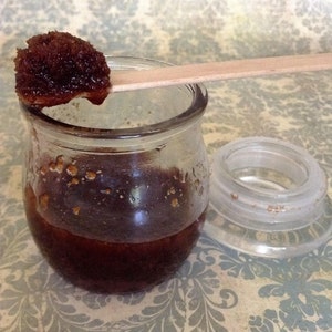 Op de afbeelding: Een glazen pot gevuld met een donkerbruine suiker scrub. Een houten stok houdt een kleine hoeveelheid van de scrub.