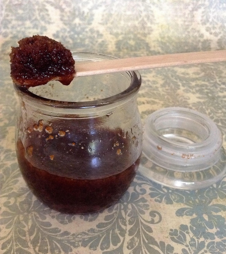 Op de afbeelding: Een glazen pot gevuld met een donkerbruine suiker scrub. Een houten stok houdt een kleine hoeveelheid van de scrub.