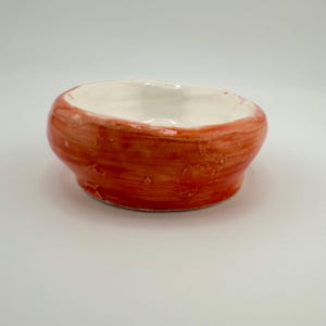 May include: A small, handmade ceramic bowl with a rounded shape. The exterior is painted with a textured, reddish-orange glaze, while the interior is white. The bowl is suitable for holding small items or as a decorative piece.