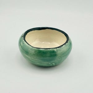 Puede incluir: Cuenco pequeño y redondo de cerámica, verde por fuera y crema por dentro. El borde es oscuro. Tiene un aspecto texturizado y pintado a mano. Probablemente se utiliza para servir o exhibir.