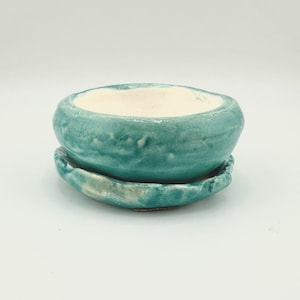 Puede incluir: Pequeña maceta de cerámica artesanal con esmalte turquesa. La maceta tiene forma redondeada y una base a juego. El interior de la maceta es de color blanco crema. Adecuada para plantas pequeñas. Diámetro aproximado de 10 cm.