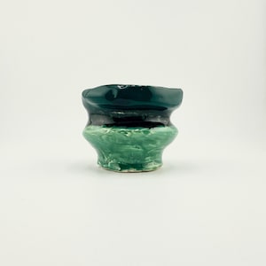 May include: A small, handcrafted ceramic vessel with a unique, layered design. The top section is a deep teal, transitioning to a dark green band, and then a textured, light green base. The vessel has a slightly irregular, organic shape.