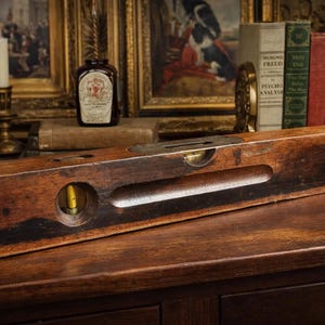 Puede incluir: Nivel de madera antiguo con detalles de latón y una ampolla de burbuja. El nivel es de color marrón oscuro con signos de desgaste, y las iniciales "H.A.L." son visibles. Descansa sobre una superficie de madera oscura, una herramienta de carpintería o construcción.
