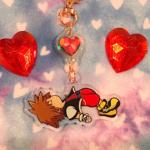 May include: A gold keychain with a red heart charm and a cartoon character charm. The character is a young person with brown hair and is wearing a black shirt and red shorts. The keychain is on a pink and blue background with white hearts.