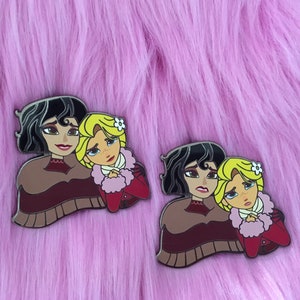 Peut inclure: Deux épingles en émail représentant des personnages de dessins animés. Les personnages sont deux femmes, l'une avec les cheveux noirs et l'autre avec les cheveux blonds. Elles portent des vêtements d'hiver et se serrent dans les bras. Les épingles sont sur un fond rose duveteux.