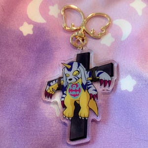 May include: A yellow and blue cartoon character with a horn is tied to a black cross. The character is a Digimon, a type of digital monster. The cross is attached to a gold keychain.
