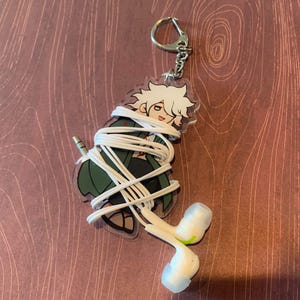 May include: A clear acrylic keyring featuring a cartoon character with spiky white hair and a green coat, wrapped in a long white cord with white wired earbuds attached at the end. The keyring includes a silver metal ring and a short chain connector.