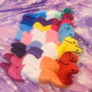Puede incluir: Una colección de coloridos gusanos de peluche dispuestos sobre una tela lavanda con un patrón celestial. Cada gusano presenta una combinación de colores diferente, incluyendo rosa, azul, amarillo y rojo, con antenas negras.