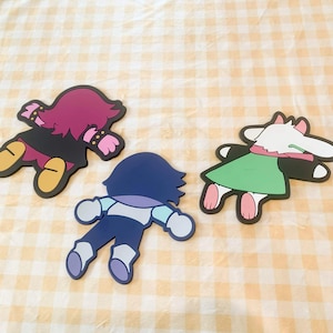 May include: Three flat, rubberized character coasters featuring stylized figures in prone positions. One figure has magenta hair and a black outfit, one is dark blue with light blue accents, and one is white with a green tunic and pink ears.