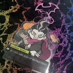 Puede incluir: Un llavero de acrílico con un personaje de dibujos animados de pelo blanco y negro, bufanda morada y vendas en un brazo. Incluye dos pequeños hámsteres naranjas. Una placa negra dice Gundham Tanaka y MY FOUR DARK DEVAS OF DESTRUCTION!!!. El llavero tiene un cierre rosa con forma de murciélago.