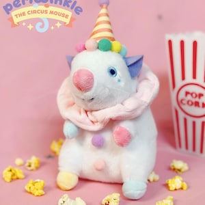 Puede incluir: Un peluche de ratón blanco con nariz, orejas y detalles rosas, que lleva un gorro de fiesta y una bufanda rosa. Palomitas de maíz esparcidas alrededor del juguete sobre una superficie rosa. El texto "Periwinkle The Circus Mouse" es visible.