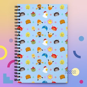 May include: A spiral-bound notebook with a light blue cover featuring a repeating pattern of cartoon characters in various colors. The characters include chickens, bears, and other whimsical designs. The notebook has a black spiral binding.