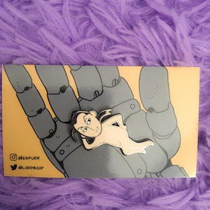 Könnte beinhalten: Anstecknadel mit einer Cartoonfigur mit schwarzen Haaren und cremefarbenem Körper in entspannter Pose, die auf einer grauen Handillustration ruht. Die Anstecknadel befindet sich auf einer gelben Karte mit Social-Media-Handles.