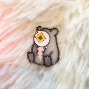 Puede incluir: Un pin de esmalte gris de un oso de dibujos animados con un ojo rosa y amarillo. El oso está sentado con las patas delante de él.