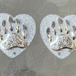 May include: A pair of silver heart-shaped earrings with a bear paw print design. The paw print is outlined in gold and has a textured surface.