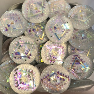 May include: A collection of iridescent, round, white craft beads. Each bead features a geometric design with a central diamond shape and surrounding triangular patterns. The beads have a glittery, textured surface and two small holes.