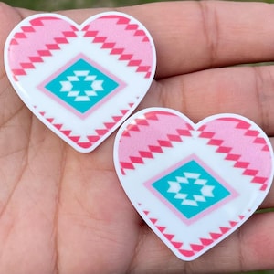 May include: Two heart-shaped decorative pieces with a Southwestern design. The hearts feature a pink and white pattern with a turquoise diamond in the center. The hearts are held in a person's hand.