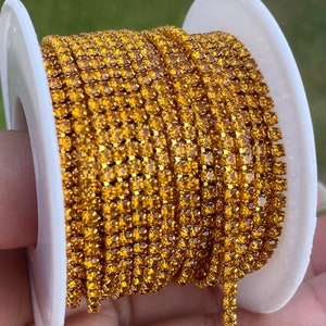 May include: A spool of gold-colored rhinestone chain. The chain is tightly wound around a white plastic spool. The rhinestones are small and closely set, creating a continuous, sparkling line. The chain is likely used for jewelry making or embellishments.