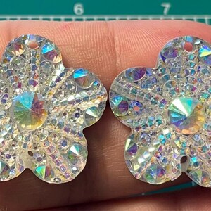 May include: Two iridescent flower-shaped embellishments with a clear background and a central iridescent stone. The embellishments are made of plastic and have a hole in the center for sewing or attaching.