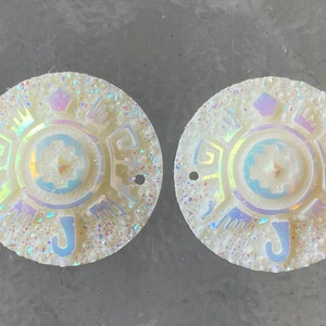 May include: Two white iridescent round decorative pieces with a geometric design and a letter J on the bottom.