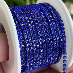 May include: A spool of blue rhinestone chain. The chain is made of small, square rhinestones that are connected together. The chain is wrapped around a white spool.