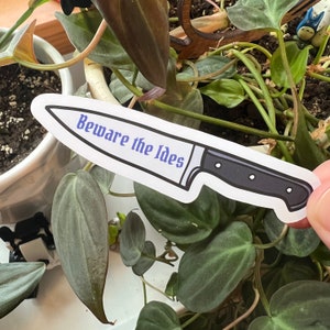 May include: A white sticker with a black and silver knife illustration. The knife has a black handle and the text "Beware the Ides" in blue lettering.