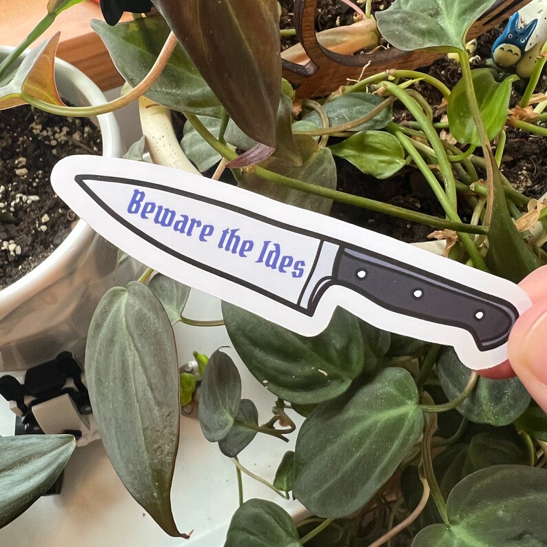 May include: A white sticker with a black and silver knife illustration. The knife has a black handle and the text "Beware the Ides" in blue lettering.