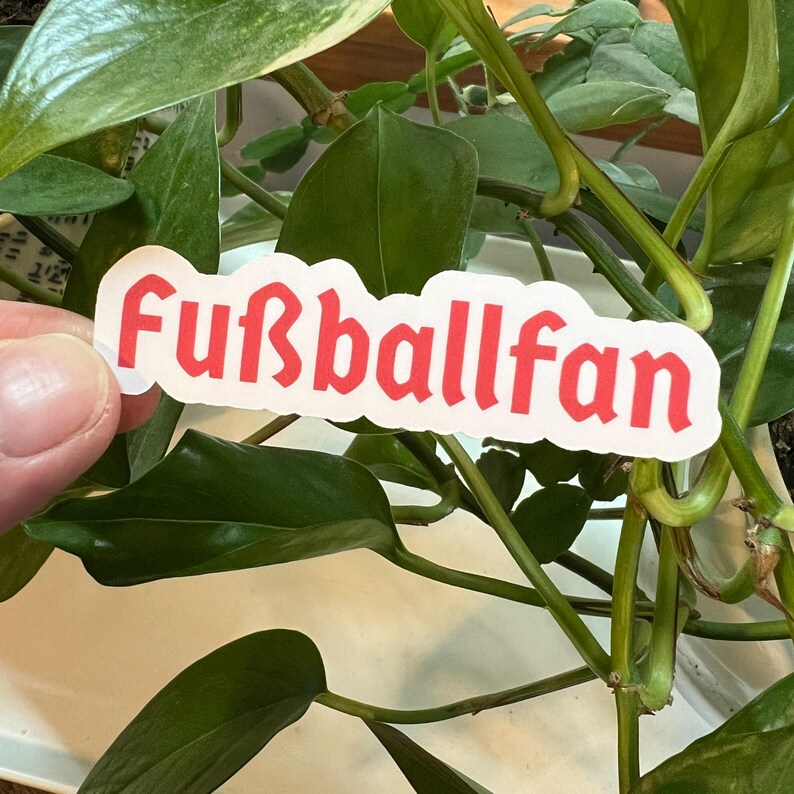 May include: A white sticker with the text "Fu&szlig;ballfan" in red, gothic-style lettering. The sticker is held by a hand against a green leafy background.