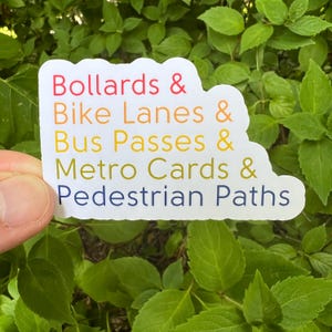 May include: A white sticker with the words "Bollards & Bike Lanes & Bus Passes & Metro Cards & Pedestrian Paths" in a rainbow gradient of colors. The sticker is held in front of a background of green leaves.