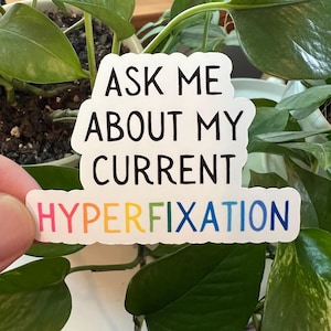 May include: White sticker with the text "Ask me about my current hyperfixation" in black. The word "hyperfixation" is in rainbow colors.