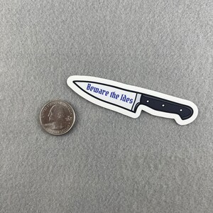 May include: A white sticker with a black and silver knife illustration. The knife has a black handle and the text "Beware the Ides" in blue.