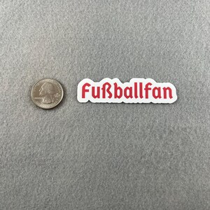 May include: White sticker with the red text "Fu&szlig;ballfan" on a gray surface.