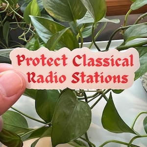 May include: A sticker with the text "Protect Classical Radio Stations" in red letters on a white background. The sticker has a wavy edge and is held in front of a background of green leaves.