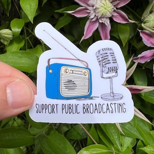 May include: A sticker featuring a blue retro radio and a vintage microphone. The text "SUPPORT PUBLIC BROADCASTING" is printed below the illustrations. The sticker is set against a backdrop of green foliage and purple flowers.