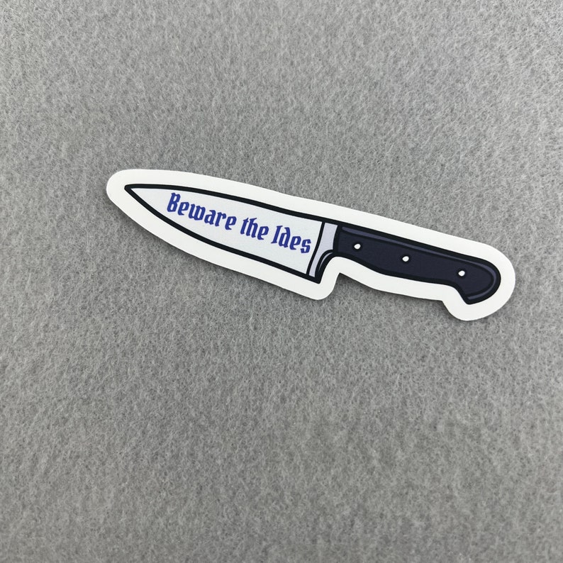 May include: A white sticker with a black and silver knife illustration. The knife has the text "Beware the Ides" written on the blade.