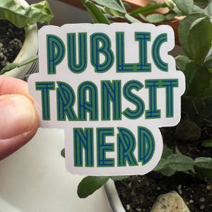 May include: A white sticker with the text "PUBLIC TRANSIT NERD" in green and blue. The sticker is held in front of a plant with green leaves. The text is in a bold, stylized font.