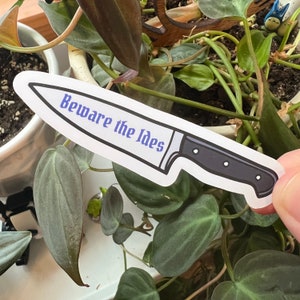 May include: A white sticker with a black and silver knife illustration. The knife has a black handle and the text "Beware the Ides" in blue.