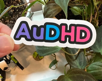 Pode incluir: Um adesivo branco com o texto "AuDHD" em letras roxas, azuis e rosas com uma contorno preto. O adesivo é segurado em frente a um fundo de plantas verdes.