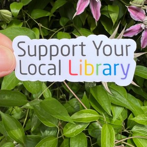 May include: A white sticker with the text "Support Your Local Library" in black letters. The word "Library" is in rainbow colors. The sticker is held in front of a background of green leaves and pink flowers.