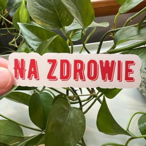 May include: A white sticker with the Polish phrase "NA ZDROWIE" in bold red letters. The sticker is rectangular with rounded corners. The background features green leaves and stems.