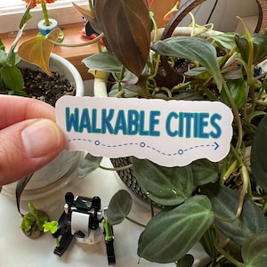 May include: A white sticker with blue and green text that reads "WALKABLE CITIES" with a dashed line and arrow. The sticker is being held by a hand in front of a potted plant.